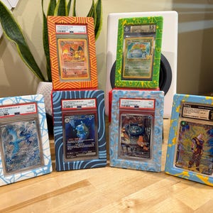 May include: Six framed collectible trading cards, featuring various characters. The frames are painted in vibrant colors, including orange, green, blue, and yellow. Each card is encased in a protective holder, showcasing the artwork and grading information.