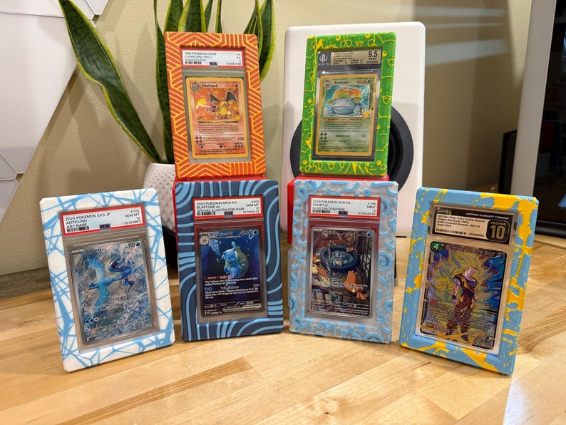 May include: Six framed collectible trading cards, featuring various characters. The frames are painted in vibrant colors, including orange, green, blue, and yellow. Each card is encased in a protective holder, showcasing the artwork and grading information.