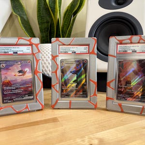 May include: Three framed Pok&eacute;mon trading cards. Each card is encased in a silver frame with a red, cracked-stone design. The cards feature images of Charizard, Charmander, and Charmeleon, with text indicating they are Illustration Rare cards.