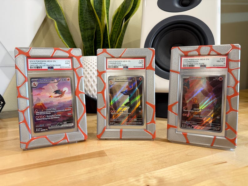May include: Three framed Pok&eacute;mon trading cards. Each card is encased in a silver frame with a red, cracked-stone design. The cards feature images of Charizard, Charmander, and Charmeleon, with text indicating they are Illustration Rare cards.