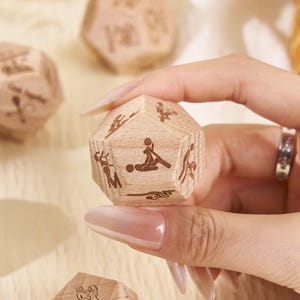 May include: Wooden dodecahedron dice with engraved illustrations of various sexual positions. The dice are light brown and appear to be made of wood. Several dice are visible, some in a woven basket. One die is held in a hand.