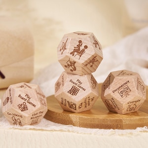 Può includere: Dadi dodecaedrici in legno con icone di attività e testo incisi. I dadi presentano opzioni come "Gioca ai giochi da tavolo", "Fai escursioni" e "Guarda film". Sono esposti su una tavola di legno.