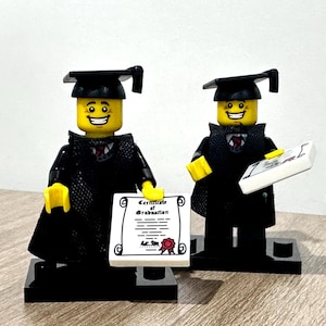 May include: Two Lego figures dressed in graduation attire. Each figure wears a black graduation gown and mortarboard cap. One figure holds a diploma with the words "Certificate of Graduation". The figures stand on black bases, with yellow hands and smiling faces.