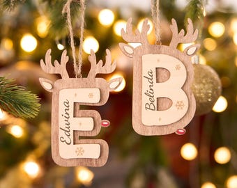 May include: Two wooden Christmas ornaments shaped like reindeer, each with a letter and a name. One ornament has the letter "E" and the name "Edwina", the other has the letter "B" and the name "Belinda". Both are hanging from twine.