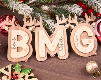 May include: Wooden Christmas ornaments in the shape of reindeer with the letters BMG and names Belinda, Michelle, and Gloria. The ornaments are light brown with snowflake details and red accents. Festive holiday decor.