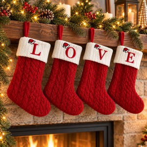 Peut inclure: Quatre chaussettes de Noël en tricot torsadé rouge avec des revers blancs suspendues en rangée. Chaque revers présente une lettre rouge brodée formant le mot LOVE et une petite icône de bonnet de Père Noël au-dessus.