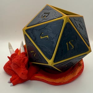 Op de afbeelding: Een rode drakenfiguur met een gouden en zwarte D20 dobbelstenenhouder op zijn rug. De dobbelstenenhouder heeft de nummers 1 tot en met 20 op zijn zijden.