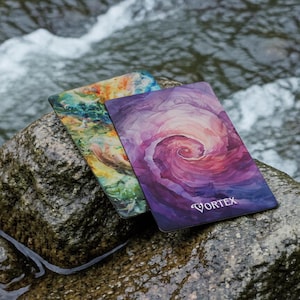 May include: Two rectangular cards with rounded corners. The front card features a purple and pink watercolour-style spiral design with the word VORTEX printed in white. The back card displays a multi-coloured abstract pattern of green, blue, and orange.