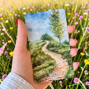 May include: A rectangular card featuring a watercolour illustration of a winding dirt path leading up a green grassy hill towards a blue sky with clouds. A single tall tree stands on the right side of the path. The word PATH is printed in white.