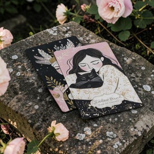 May include: Two decorative cards with illustrations of a woman and a black cat, set against a black and pink background. The cards are resting on a stone surface, surrounded by pink roses. The text "Shadow Mind" is visible.