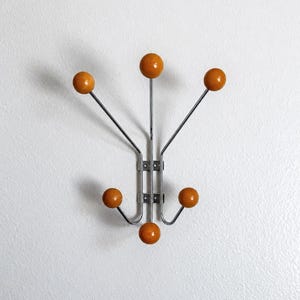 Op de afbeelding: Een wandkapstok met een verticaal frame van verchroomd metaal en zes uitstekende armen. Elke arm eindigt in een gladde, ronde houten bol in de kleur mosterdgeel. De centrale structuur bevat twee rechthoekige montageplaten met schroefgaten.