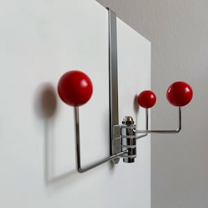 May include: A chrome over-the-door hook rack featuring three distinct arms, each tipped with a smooth, bright red spherical knob. The metal structure includes a flat vertical bracket designed to hang securely over the top edge of a door.
