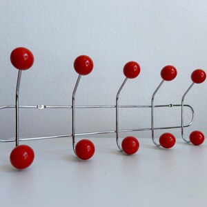 May include: A chrome metal wall-mounted coat rack with eight bright red ball-shaped hooks. The hooks are evenly spaced along a horizontal bar, creating a mid-century modern design. The rack is displayed against a white background.