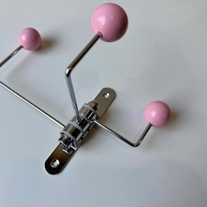 May include: A modern wall hook with a silver metal frame and three pink spherical knobs. The hook is mounted on a rectangular metal plate with screw holes. The design is minimalist and functional, suitable for hanging items.