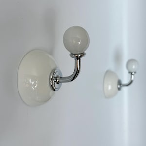 May include: Two wall-mounted hooks featuring a polished chrome metal arm and a smooth, glossy white ceramic base. Each hook is topped with a spherical white ceramic knob, providing a functional design for hanging items on a flat surface.