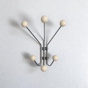 May include: A wall-mounted coat rack featuring a vertical chrome metal frame with six extending arms. Each arm terminates in a smooth, round, cream-colored ball hook. The structure is symmetrical, with three hooks pointing upward and three hooks pointing downward.