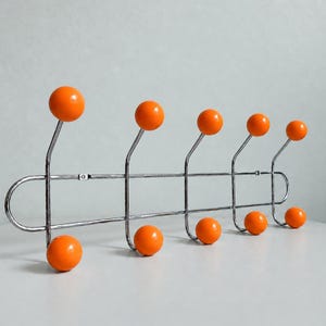 May include: A wall-mounted coat rack featuring a horizontal chrome metal frame with five upward-curving hooks and five downward-curving hooks. Each hook is tipped with a smooth, round, bright orange ball for hanging garments and accessories.