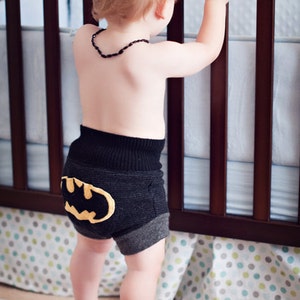 Może przedstawiać: Małe dziecko w czarnych i szarych spodenkach z ż&oacute;łtym logo Batmana z tyłu. Dziecko stoi przed ł&oacute;żeczkiem z białą i niebieską wzorzystą pościelą.