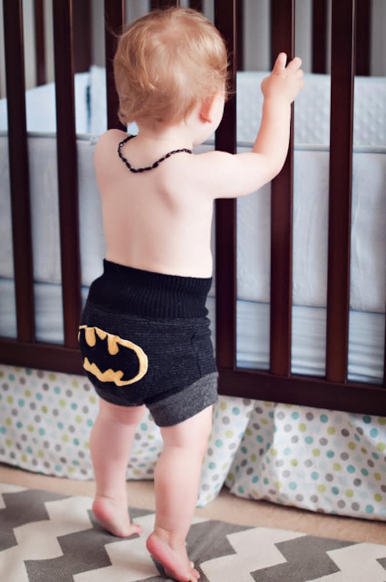 Może przedstawiać: Małe dziecko w czarnych i szarych spodenkach z ż&oacute;łtym logo Batmana z tyłu. Dziecko stoi przed ł&oacute;żeczkiem z białą i niebieską wzorzystą pościelą.