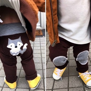 May include: A child wearing brown pants with a wolf applique on the back and gray knee pads. The child is also wearing a white long-sleeved shirt and a brown jacket. The child is wearing yellow shoes.