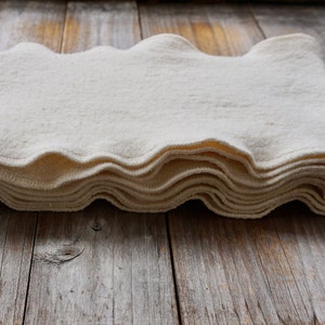 May include: A stack of white cotton fabric squares with scalloped edges. The fabric is folded and stacked on a wooden surface.