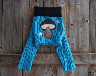 May include: Bright blue cable knit leggings featuring a black waistband and a central brown and white mushroom appliqué. Two small white star appliqués are positioned on either side of the mushroom, adding detail to the textured blue fabric.