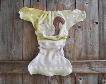 May include: A cloth diaper featuring a light yellow and white speckled pattern with a central applique of a grey squirrel with a brown tail. The garment includes multiple white plastic snap buttons along the waist and front panel for adjustment.