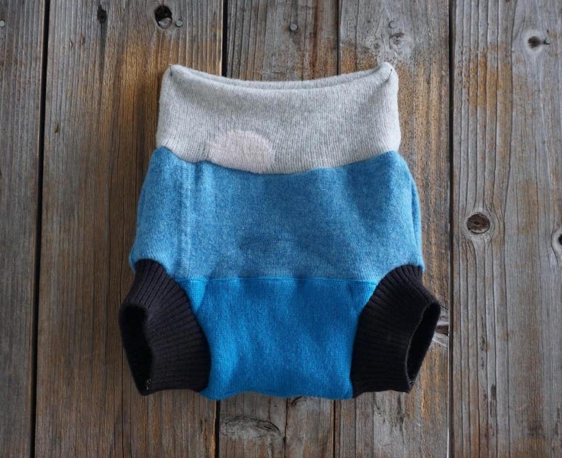 May include: A multi-colored baby diaper with a gray waistband, a blue midsection, and black leg cuffs. The diaper is made of soft fabric and is designed for comfort and absorbency. The diaper is displayed on a wooden surface.