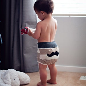 Może przedstawiać: Małe dziecko w paskowanej pieluszce z czarnym wzorem wąs&oacute;w z tyłu. Dziecko trzyma czerwony plastikowy samoch&oacute;d w ręku.