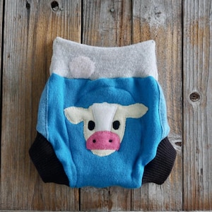 May include: A baby diaper cover with a blue body, gray waistband, and black trim. The front features a white and pink cow face applique. The diaper cover is made of soft fabric and is designed for comfort.