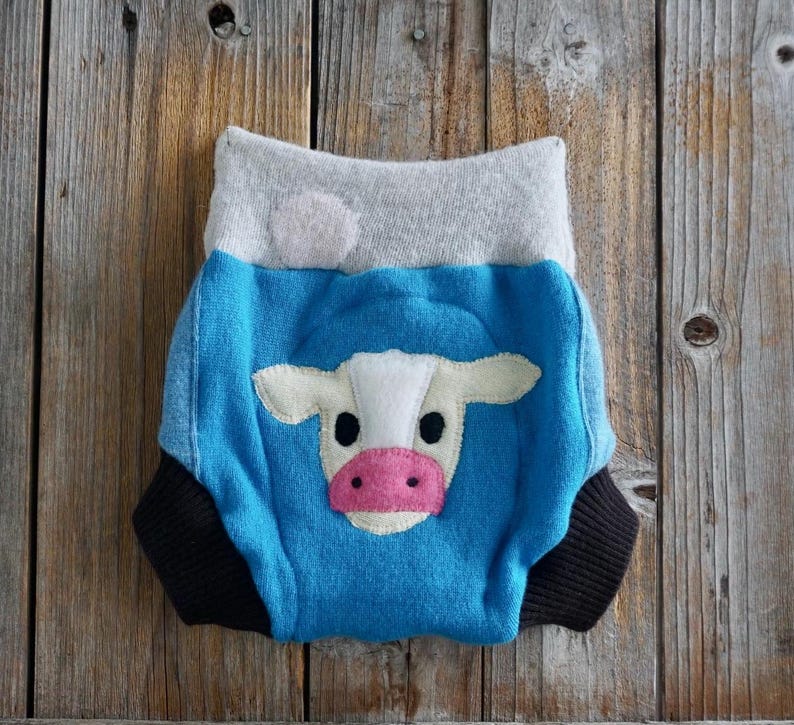 May include: A baby diaper cover with a blue body, gray waistband, and black trim. The front features a white and pink cow face applique. The diaper cover is made of soft fabric and is designed for comfort.