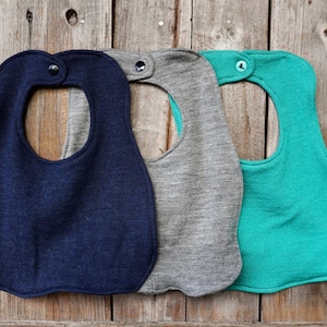 May include: Three knitted baby bibs in navy blue, gray, and teal. Each bib has a round opening for the neck and a button closure.