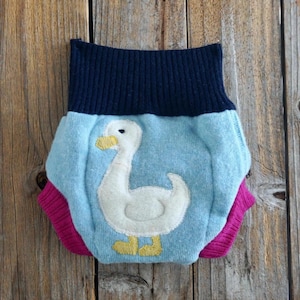 Puede incluir: Una braguita de pañal para bebé hecha de tela azul, rosa y azul marino. La braguita presenta una aplicación de ganso blanco con detalles amarillos. La braguita es de material suave.