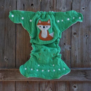May include: A green cable knit cloth diaper featuring a centered felt fox appliqué in orange, white, and black. The diaper includes white snap closures along the wings and front panel, with a textured knit pattern throughout the fabric surface.
