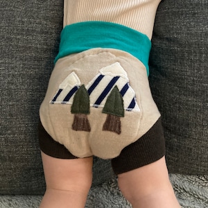 Może przedstawiać: Beżowe spodenki dla niemowląt z brązowym pasem i aplikacją przedstawiającą g&oacute;rę i drzewa. Aplikacja przedstawia g&oacute;rę w pasy niebieskie i białe z zielonymi drzewami i brązowymi pniami drzew.