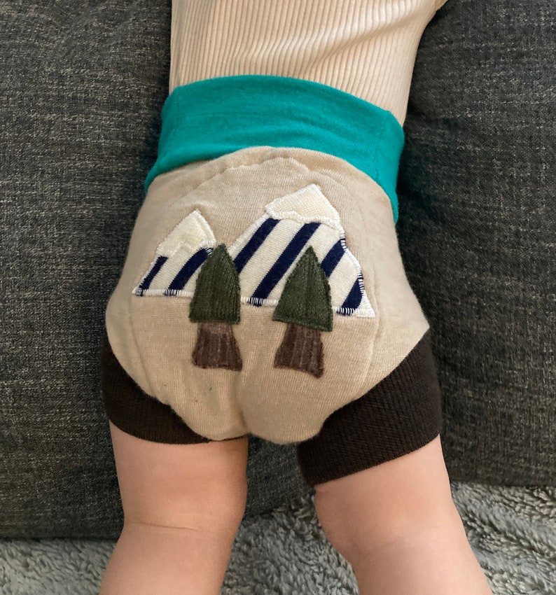 Może przedstawiać: Beżowe spodenki dla niemowląt z brązowym pasem i aplikacją przedstawiającą g&oacute;rę i drzewa. Aplikacja przedstawia g&oacute;rę w pasy niebieskie i białe z zielonymi drzewami i brązowymi pniami drzew.