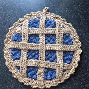 May include: A round, crocheted potholder designed to resemble a blueberry pie. It features a textured blue centre with a raised, beige lattice crust pattern and a scalloped beige border. A small loop is attached to the top for hanging.