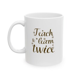 May include: White ceramic mug with a curved handle. The mug features the text "Teach to learn twice" in a brown, cursive font with snowflake and star accents. A simple, everyday coffee mug.