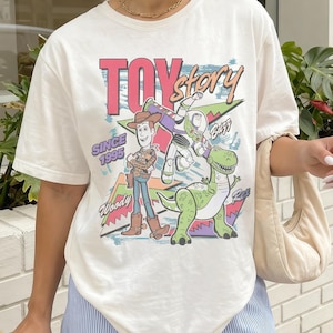 May include: Cream-colored t-shirt featuring a colorful graphic of Toy Story characters Woody, Buzz Lightyear, and Rex. The text "TOY STORY" is in pink and yellow, with "SINCE 1995" below. The shirt has short sleeves and a relaxed fit.