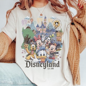 May include: A cream-colored t-shirt featuring a colorful graphic of Disney characters including Mickey Mouse, Minnie Mouse, Donald Duck, Goofy, and Pluto in front of a castle, with the text Disneyland Est. 1955 printed below the characters.