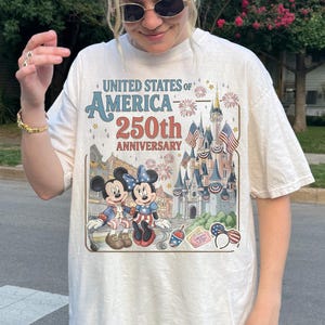 May include: Cream-colored t-shirt featuring a graphic celebrating the 250th anniversary of the United States of America. The design includes Mickey and Minnie Mouse, a castle, fireworks, and patriotic imagery. The text "United States of America 250th Anniversary" is prominently displayed.