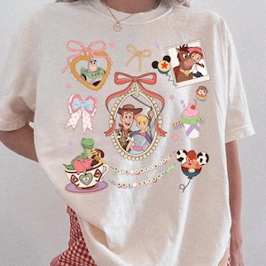 May include: Cream-coloured t-shirt with colourful cartoon graphics of Toy Story characters. The design includes Buzz Lightyear, Woody, Bo Peep, and other characters, along with the phrase "to infinity and beyond."