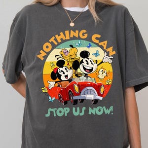 May include: Dark gray t-shirt featuring a colorful graphic of Mickey and Minnie Mouse in a red car, with Pluto and other characters. The text "NOTHING CAN STOP US NOW!" is printed on the shirt.