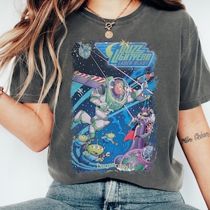 K&ouml;nnte beinhalten: Dunkelgraues T-Shirt mit einer farbenfrohen Grafik von Buzz Lightyear im Weltraum, mit dem Text "Buzz Lightyear Laser Blast" und "Discoveryland". Das Design umfasst Aliens und einen lila Schurken.