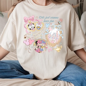 May include: Cream-colored t-shirt featuring a colorful graphic with Minnie Mouse, Daisy Duck, and a castle. The design includes pink bows, stars, and the text "Girls just wanna have fun."