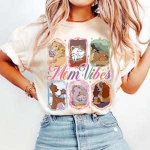 May include: Cream-colored t-shirt featuring the text "Mom Vibes" in a colorful script. The shirt displays framed illustrations of animated mothers and their children. The design includes images of lions, elephants, and dogs.