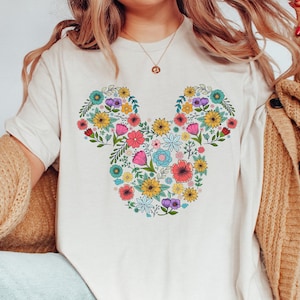K&ouml;nnte beinhalten: Cremefarbenes T-Shirt mit einem floralen Mickey-Mouse-Design. Das Design besteht aus verschiedenen bunten Blumen, darunter rosa, gelbe, blaue und lila, die in Form von Mickey Mouses Kopf und Ohren angeordnet sind. Das Shirt ist ein l&auml;ssiger, bequemer Stil.