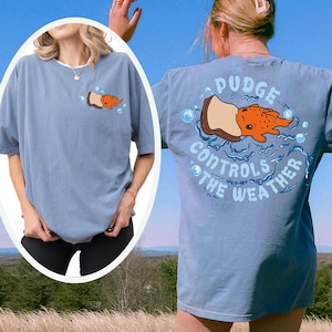 May include: A light blue t-shirt with a graphic of a cartoon fish on a piece of bread, with the text "Pudge Controls the Weather". The shirt has short sleeves and a relaxed fit. The design is on the front and back of the shirt.