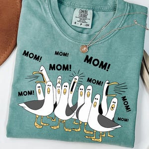 May include: A teal Comfort Colors t-shirt featuring a graphic of cartoon seagulls with the word "MOM!" repeated around them. A silver necklace with a pendant is draped on the shirt.