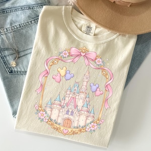 May include: Cream-colored t-shirt featuring a fairytale castle design within a pink ribbon frame, adorned with Mickey Mouse balloons and floral accents. The shirt has the text "COMFORT COLORS" on the tag. The shirt is folded on top of blue jeans.